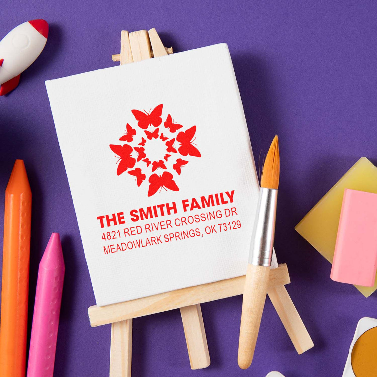 Personalized butterfly family address rubber stamp with custom name and mailing address, displayed on a canvas easel with colorful art supplies.