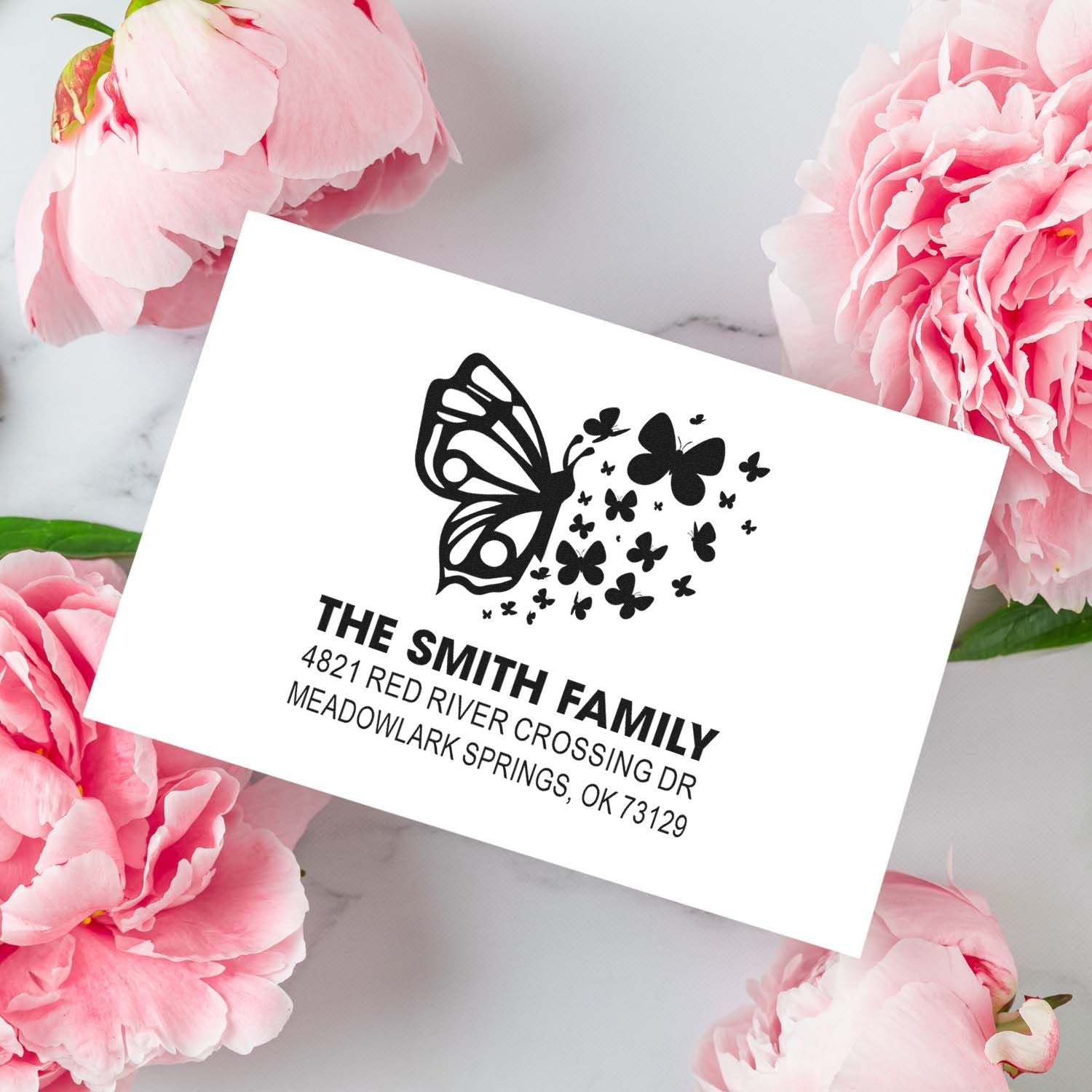Custom butterfly family return address rubber stamp with personalized name and address, elegant monogram-style design on a floral flat lay background.