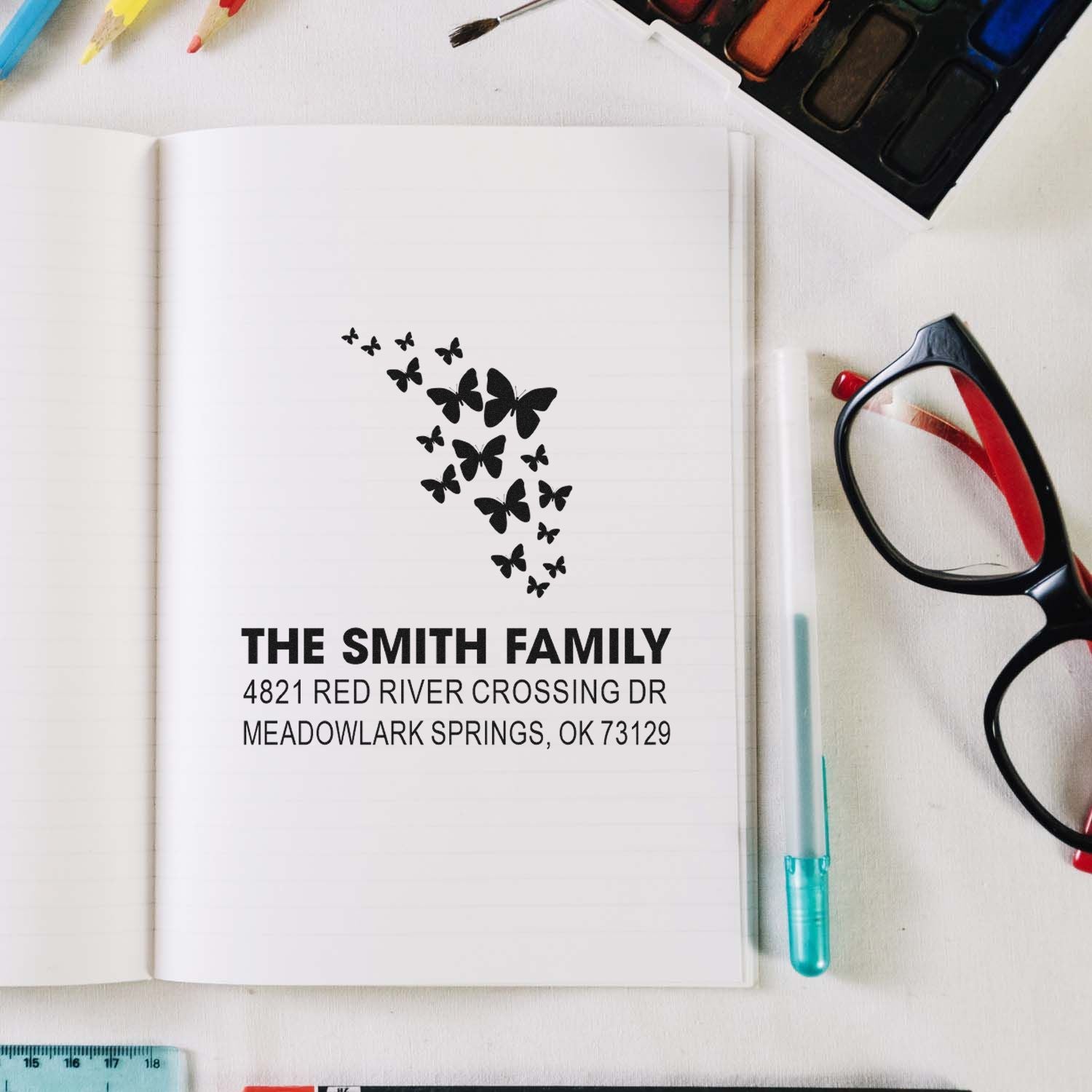 Custom butterfly family address rubber stamp with personalized name and address, shown stamped in an open notebook on a stationery desk.