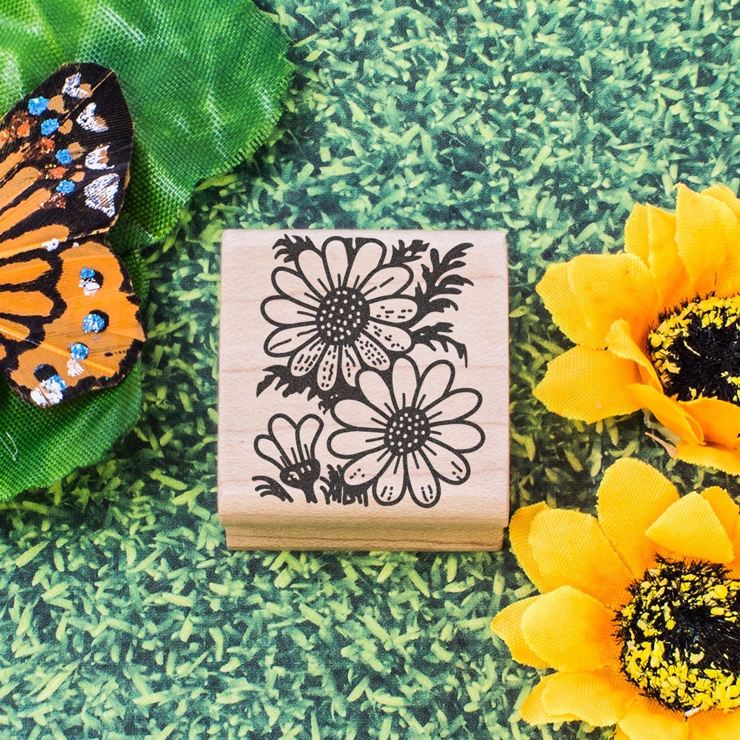 Floral rubber stamp with black daisy design on a wooden block, surrounded by sunflower decorations and a monarch butterfly wing on a green grass background.