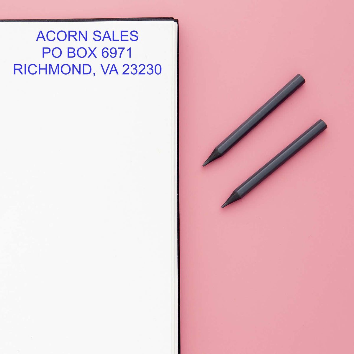 Open notebook with ACORN SALES address and two black pencils on pink background