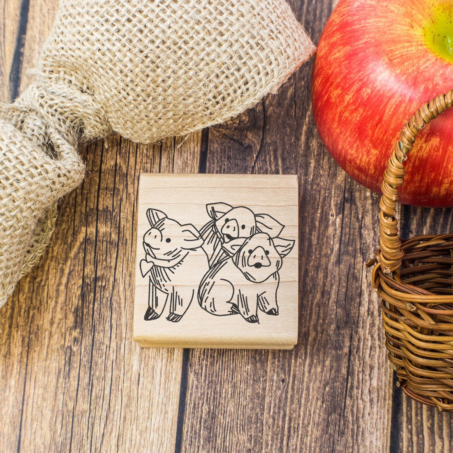 Wooden stamp with three pigs illustration on rustic table beside apple, burlap, and wicker basket.