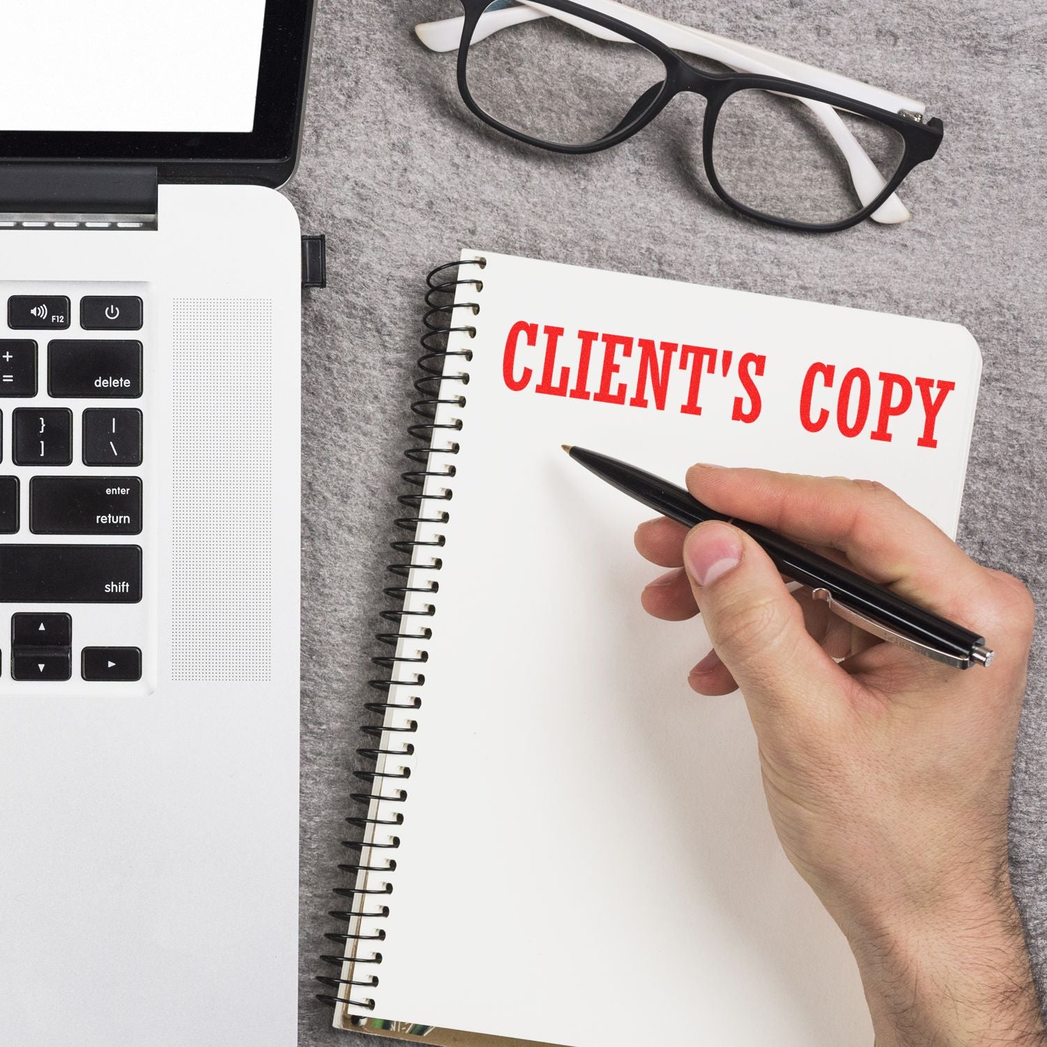 Hand writing CLIENTS COPY in notebook with pen on desk beside laptop and eyeglasses, showing document copying for clients.