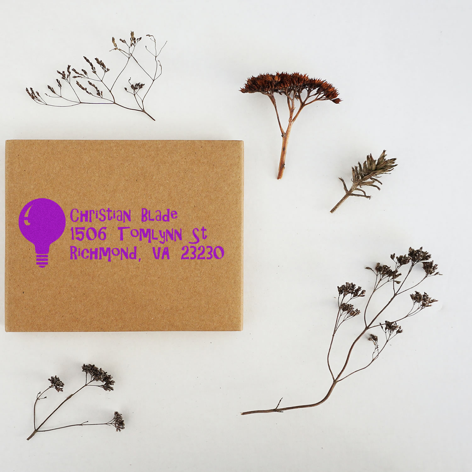 Custom address rubber stamp on kraft box with purple light bulb logo and return address, styled with dried botanicals.