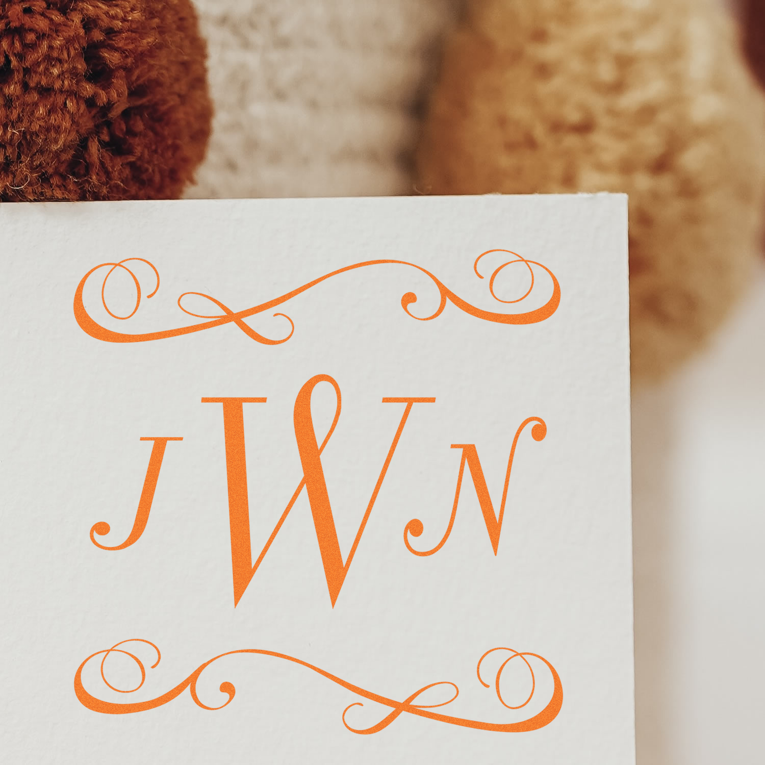 Custom monogram rubber stamp with ornate orange initials JW and decorative flourishes on white paper.