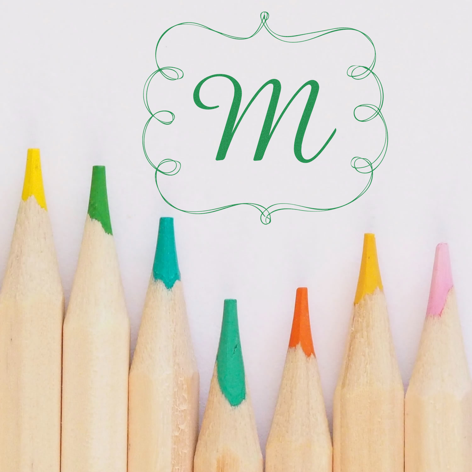 Custom monogram rubber stamp with decorative green letter M and ornate frame over colored pencils.
