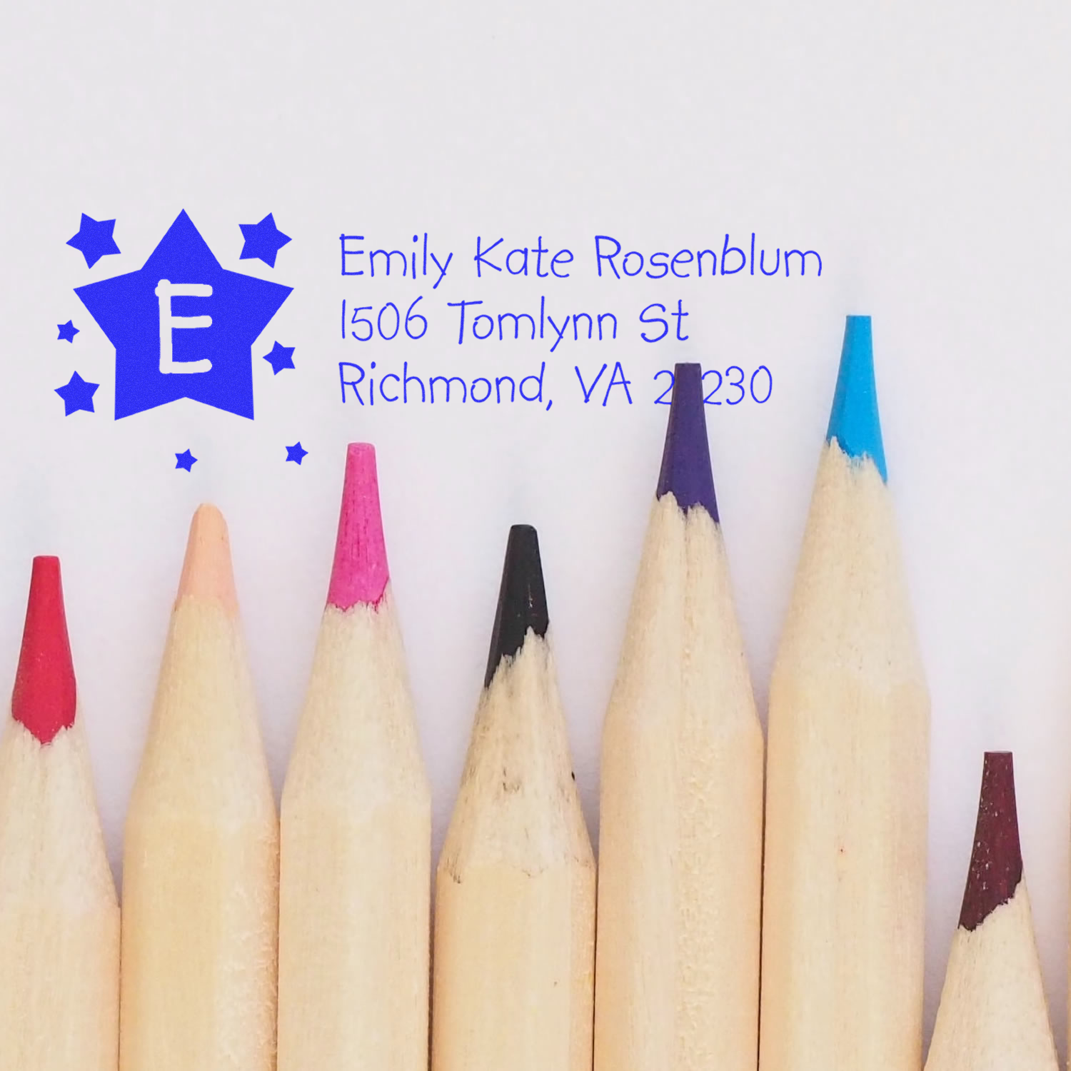 Custom return address rubber stamp with monogram E and blue imprint, shown with colored pencils for design preview.