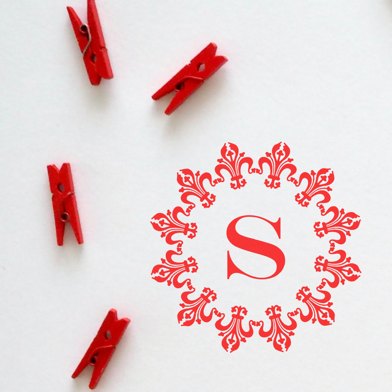 Red clothespins framing a decorative floral monogram S on a white background for craft and branding design.