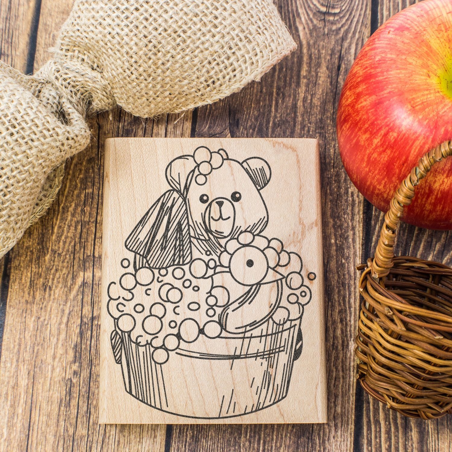 Wooden bear bath stamp with bubbles on rustic wood background, beside burlap fabric, red apple, and wicker basket.