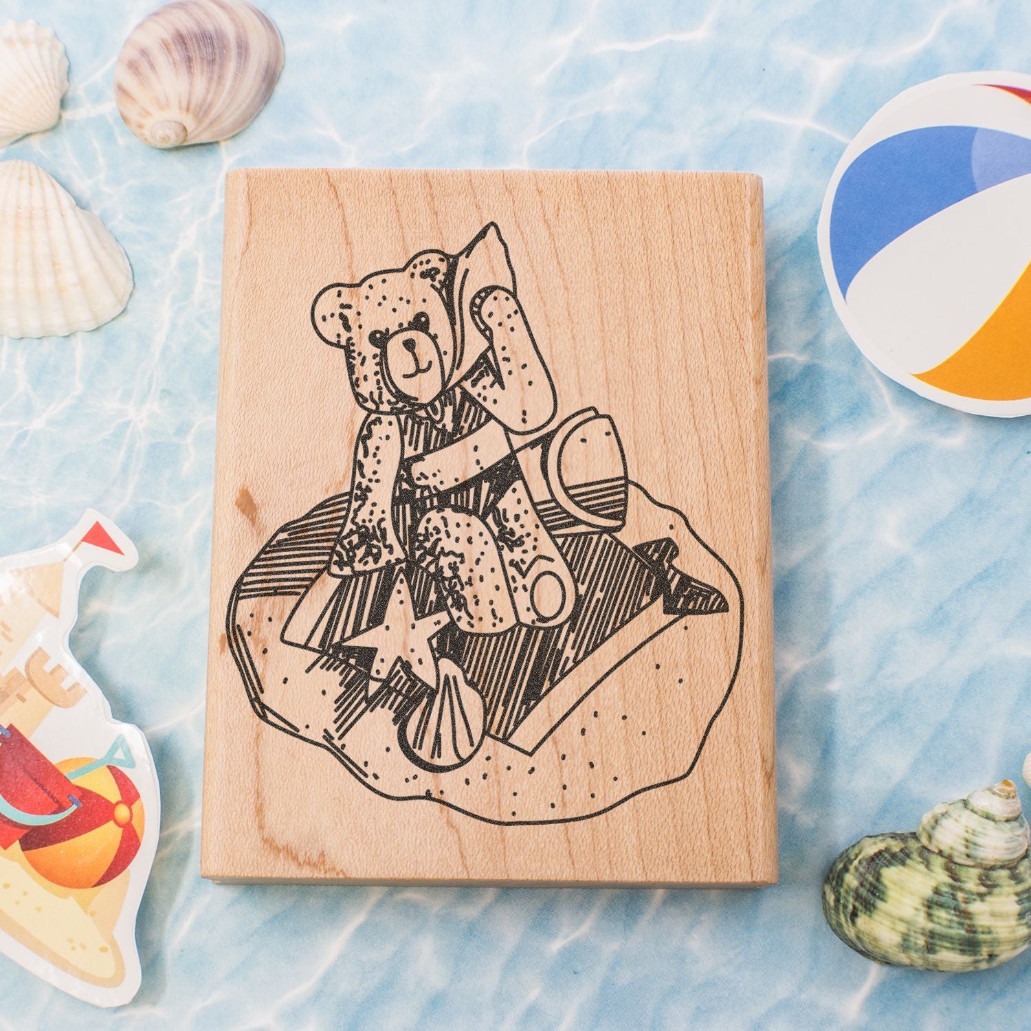 Wooden bear stamp block with beach-themed craft supplies, seashells, and colorful summer stickers on a blue water background.