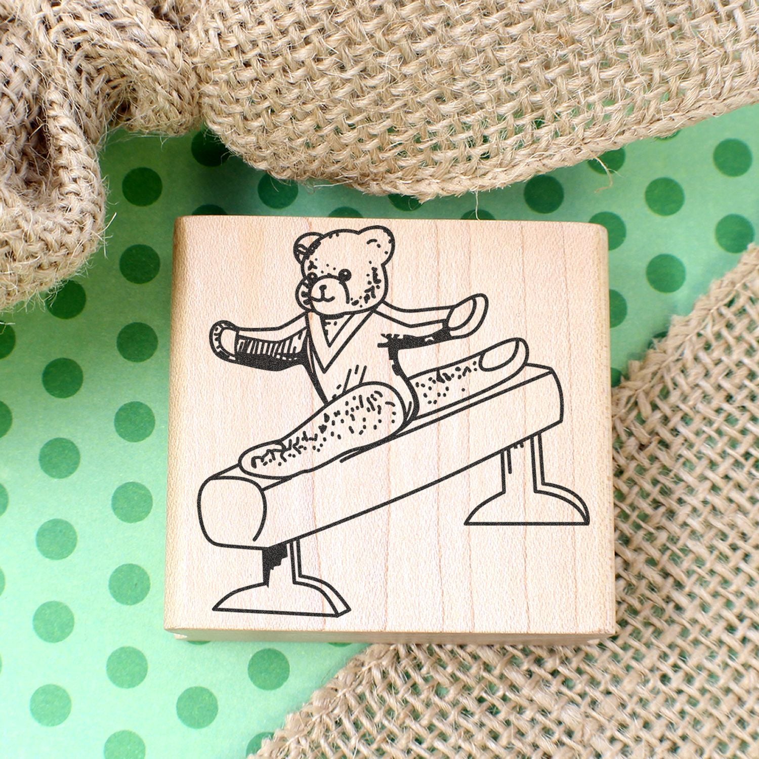 Wooden rubber stamp with bear gymnast on pommel horse, on green polka dot background with burlap texture