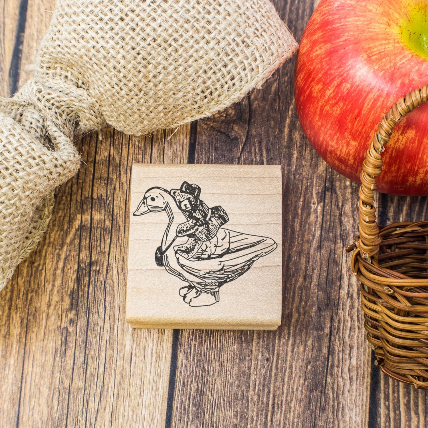 Rustic wooden rubber stamp with duck illustration, burlap fabric, red apple, and wicker basket on a wood table.