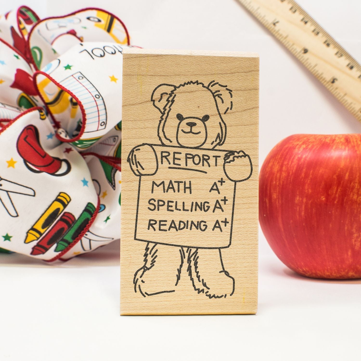 Wooden school stamp with bear holding report card showing A+ in math, spelling, and reading, surrounded by apple, ruler, and backpack.