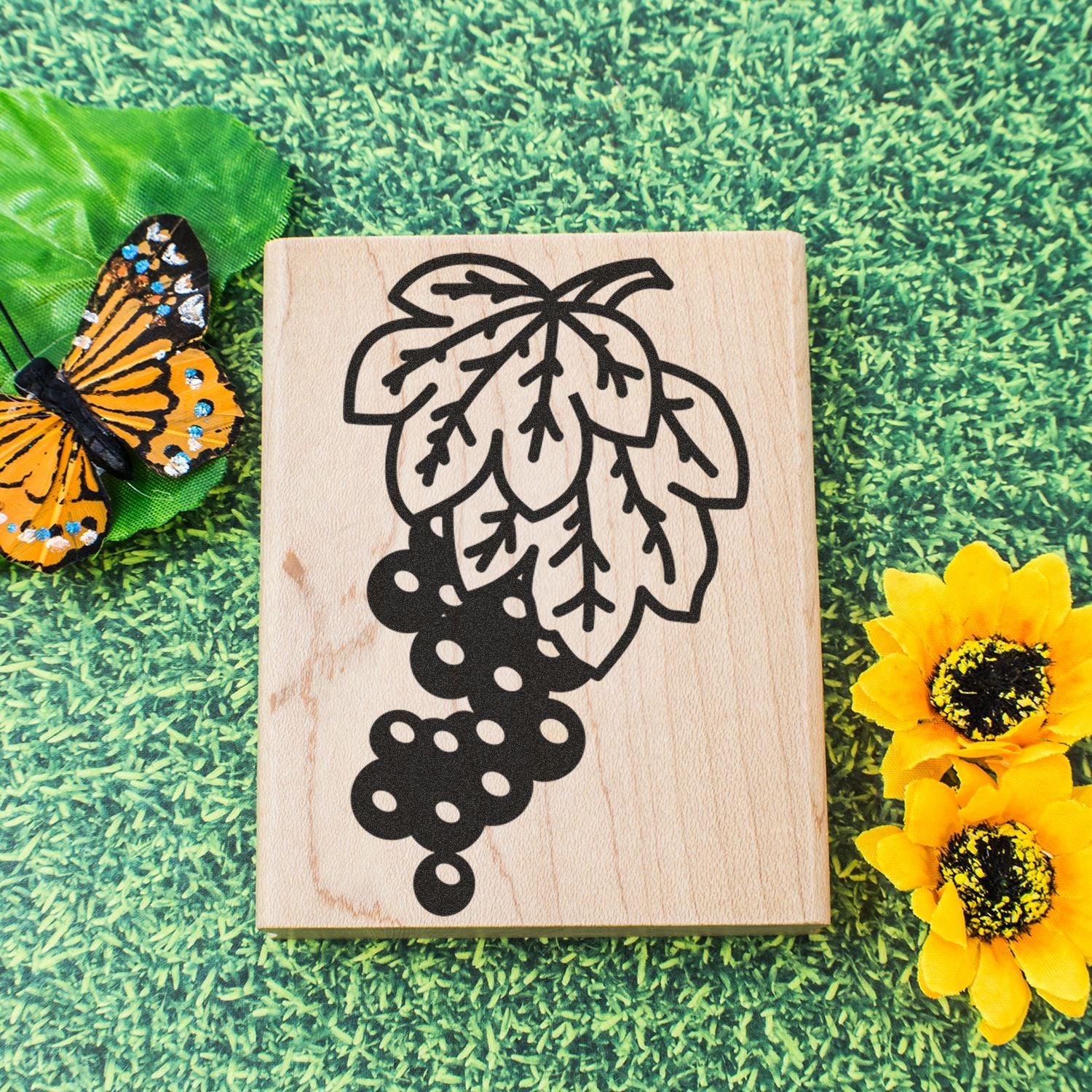 Wooden block with hop plant design, butterfly, and yellow flowers on green grass background.