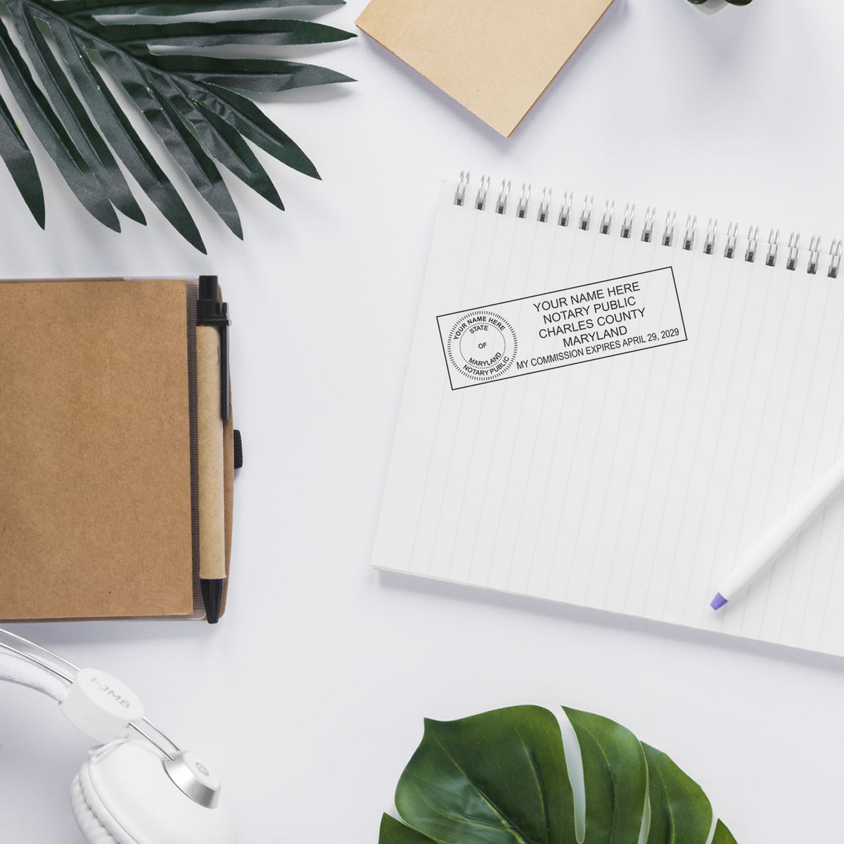 Flat lay office desk with notary public stamp on a spiral notebook, pen, notebook, headphones, and green leaves on a white background.