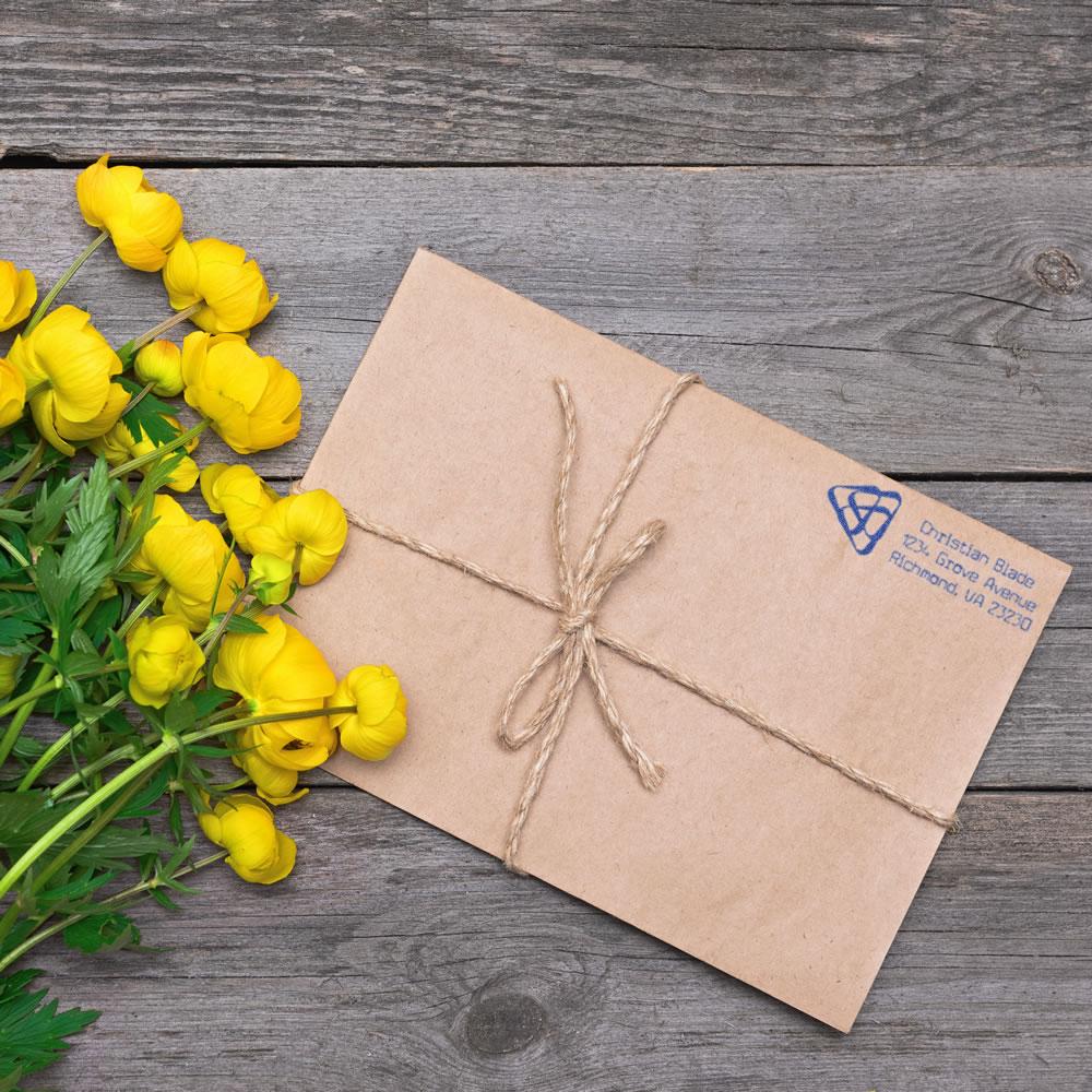 Custom return address rubber stamp on kraft paper envelope tied with twine, styled with yellow flowers on rustic wood.