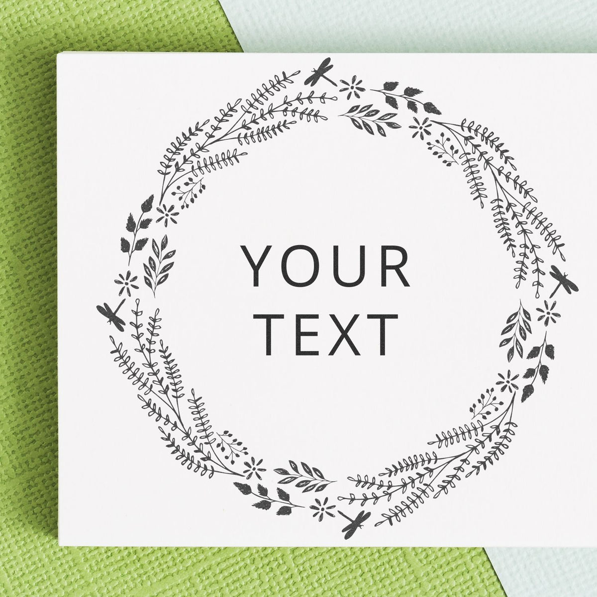 Minimalist floral wreath greeting card mockup with YOUR TEXT on a white square card over a green textured background.