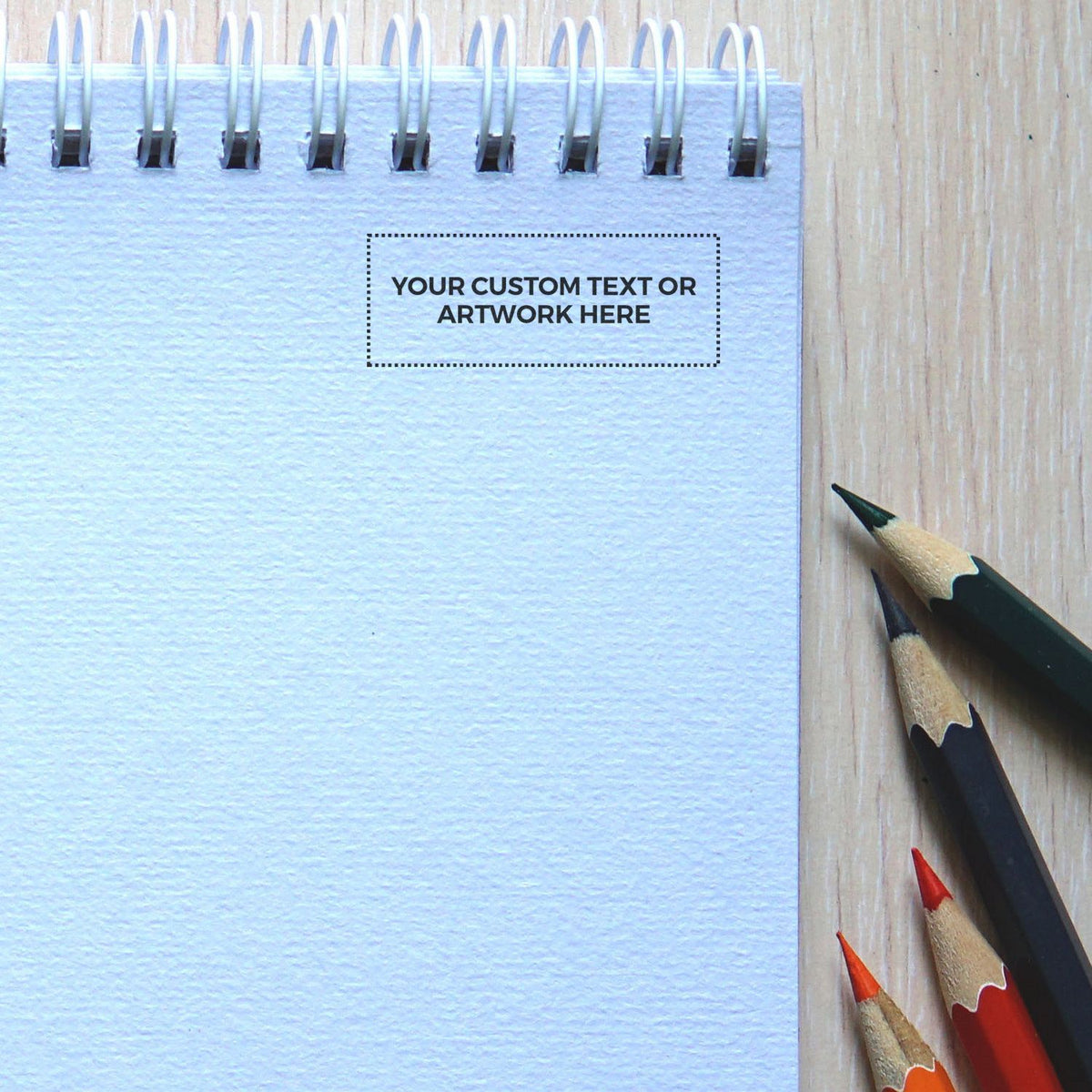 Blank spiral notebook with colored pencils on a wooden desk, customizable text and artwork template mockup.