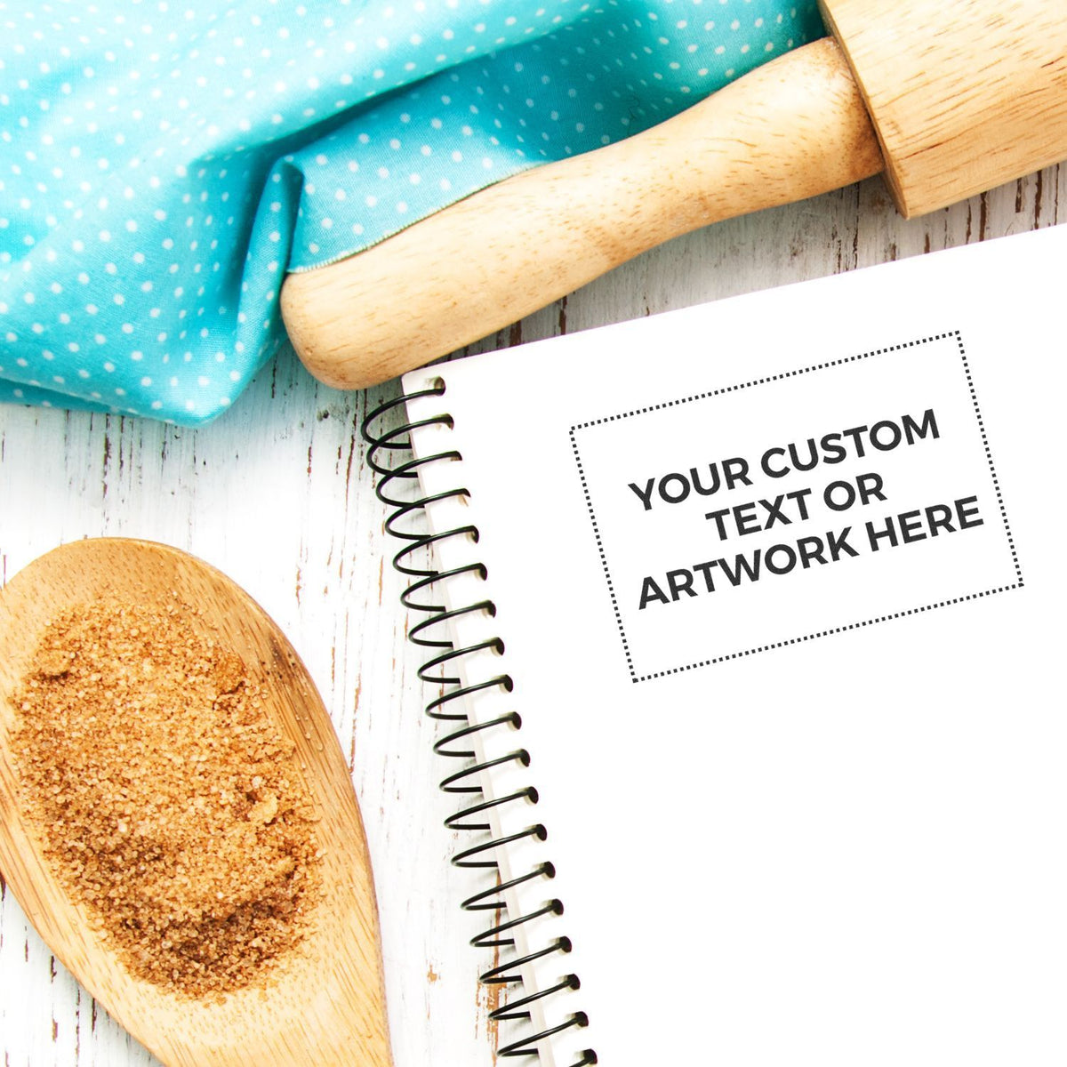 Flat lay mockup of a spiral notebook with custom text placeholder, wooden spoon, and blue polka dot fabric on a rustic white background.