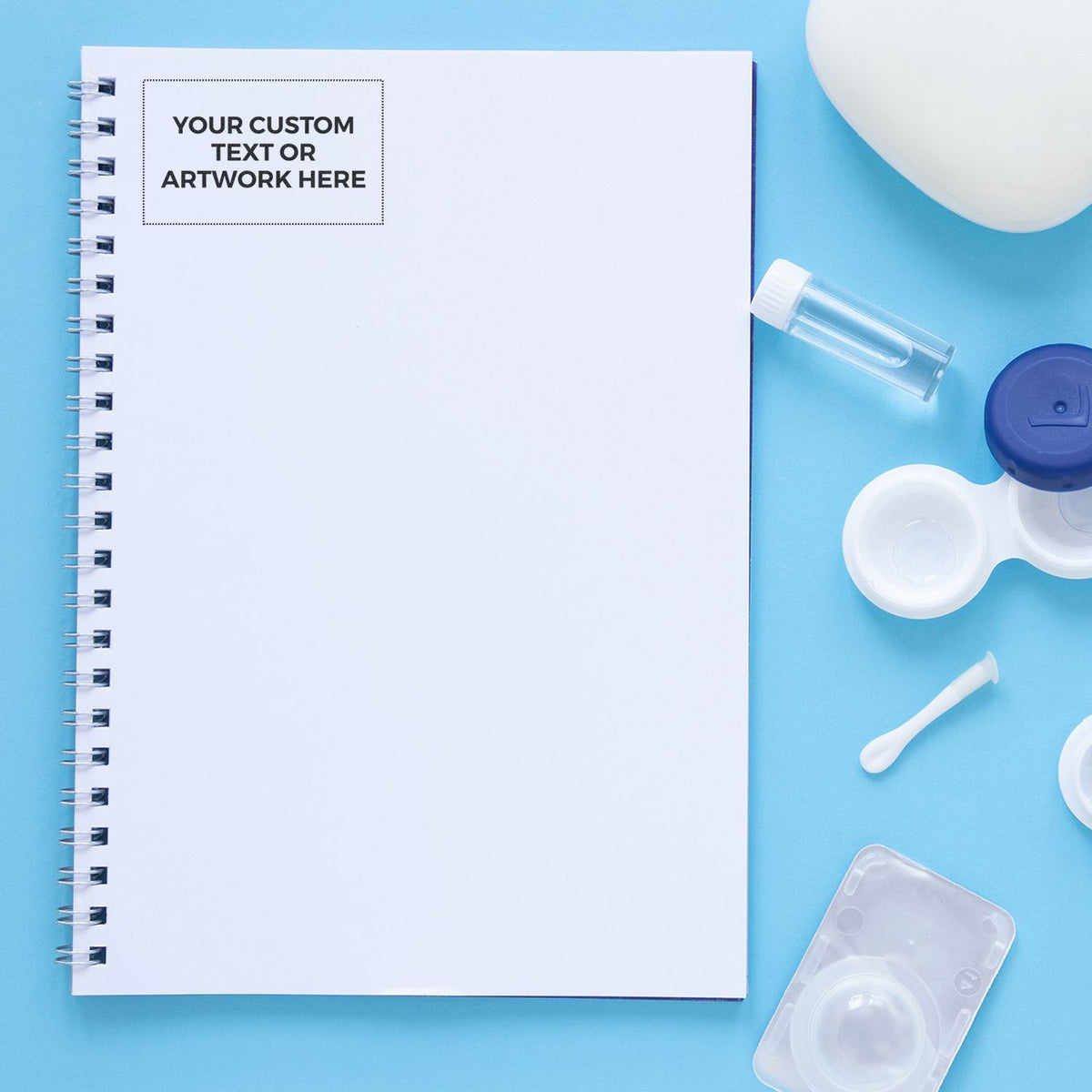Blank spiral notebook mockup with contact lens case, solution bottle, and applicator on blue background for custom branding.
