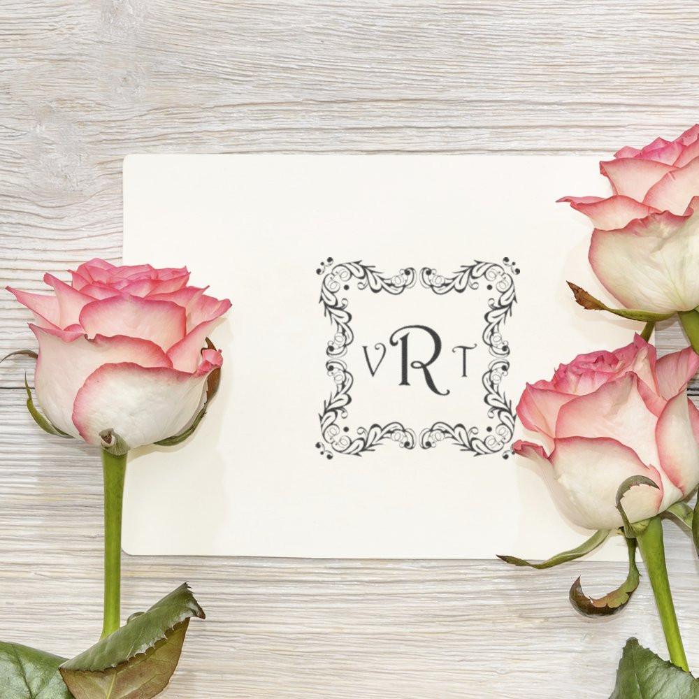 Ornate custom monogram rubber stamp with floral border on white card, styled with pink roses for wedding stationery and branding.