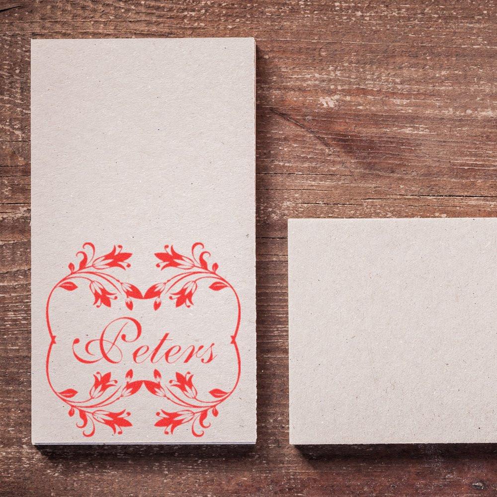 Custom red floral rubber stamp with the name Peters on cardstock, shown beside a blank card on a rustic wooden background.