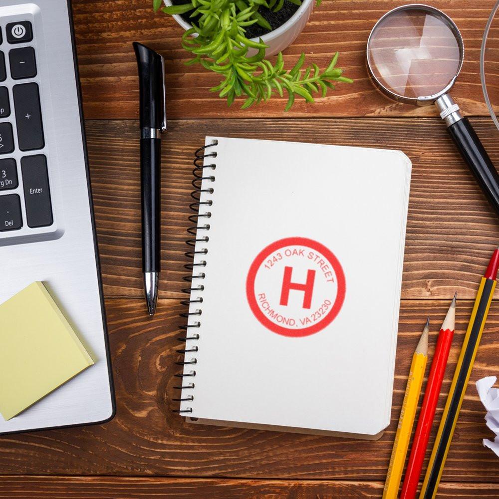 Notebook with red brand stamp and address text on wooden desk, viewed with magnifying glass, pens, and laptop keyboard for business identity.