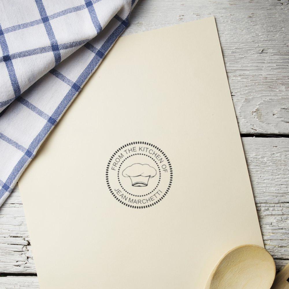 Custom rubber stamp impression on cream stationery reading "From the Kitchen of Jean Marchetti" with chef hat logo and rustic kitchen styling.