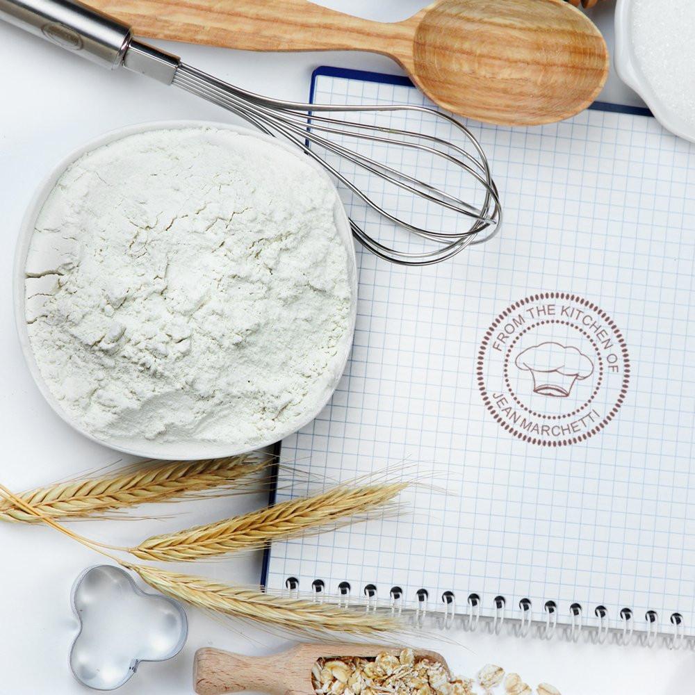 Custom bakery rubber stamp logo on a recipe notebook, surrounded by flour, whisk, and baking tools for kitchen branding.