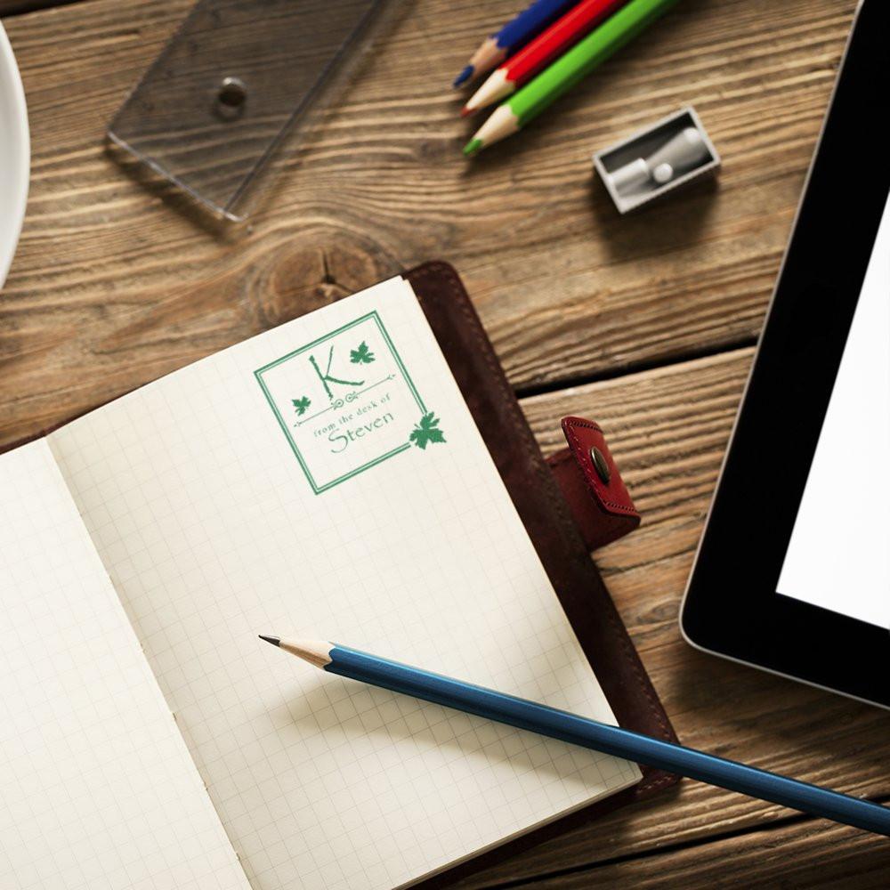 Custom rubber stamp in a notebook reading From the Desk of Steven, shown on a desk with pencils and a tablet.