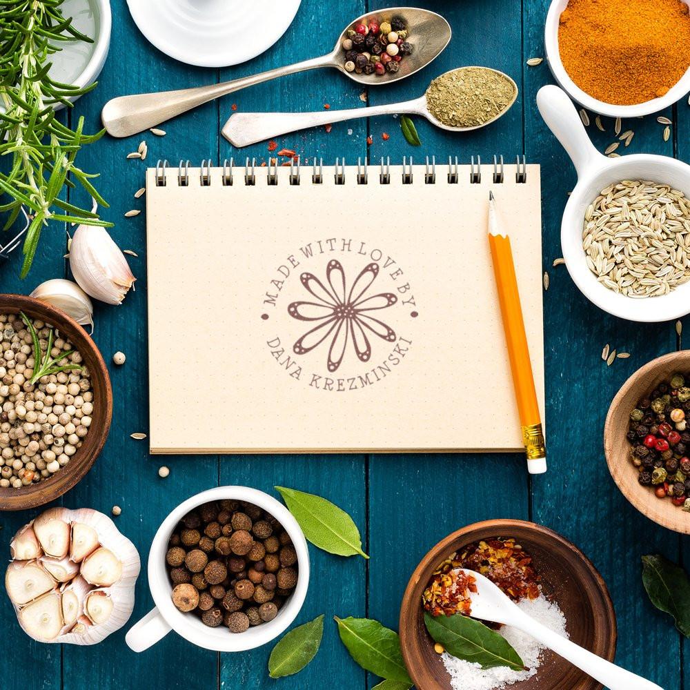 Custom rubber stamp imprint on a recipe notebook, framed by spices and herbs for handmade kitchen branding.