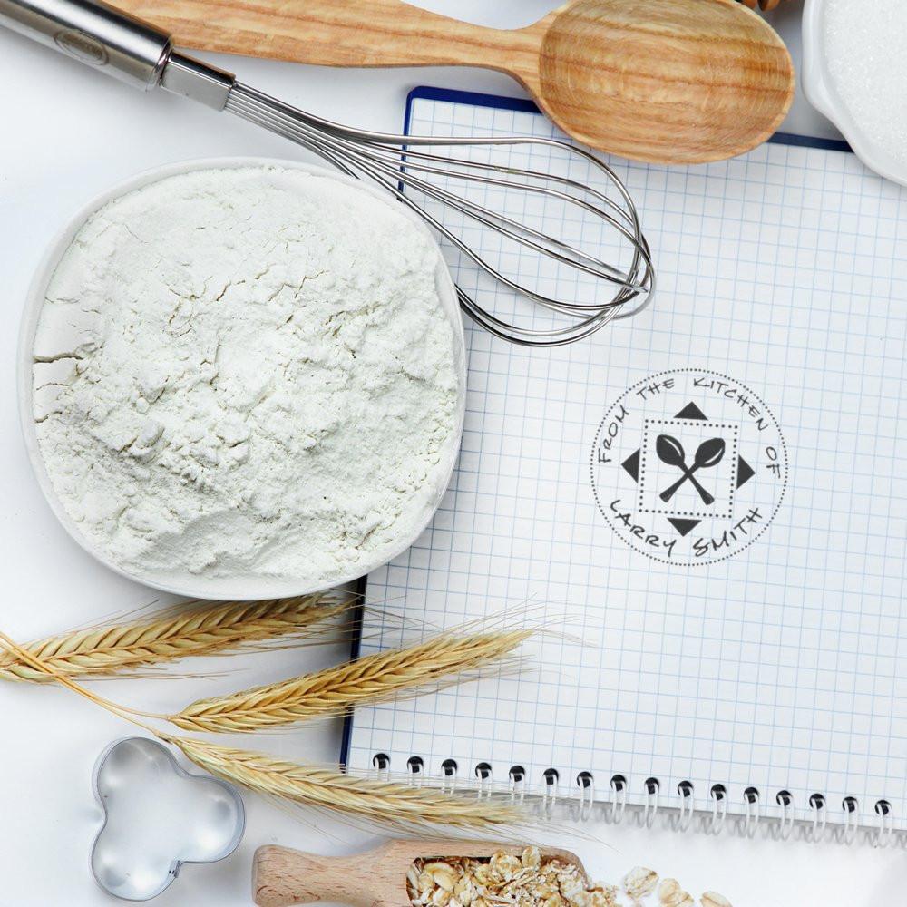 Custom rubber stamp for homemade baking recipes on a kitchen notebook, styled with flour, whisk, spoon, and wheat.