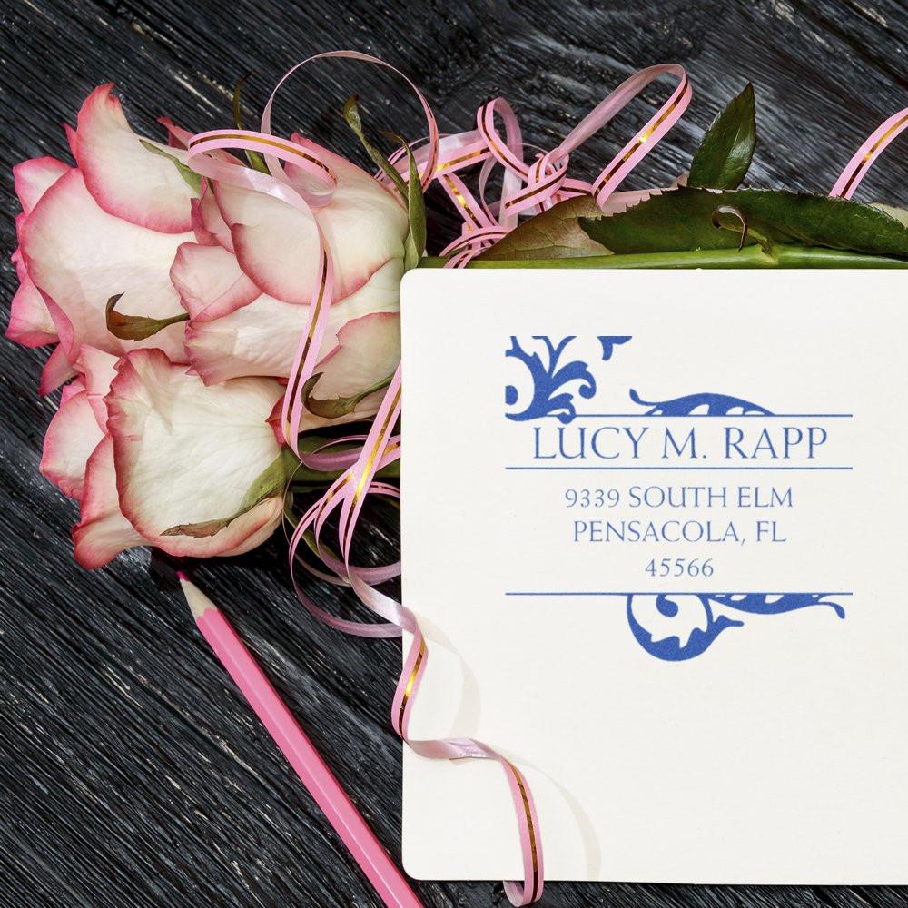 Custom return address rubber stamp with floral flourish, shown on wedding stationery with pink roses and ribbon.