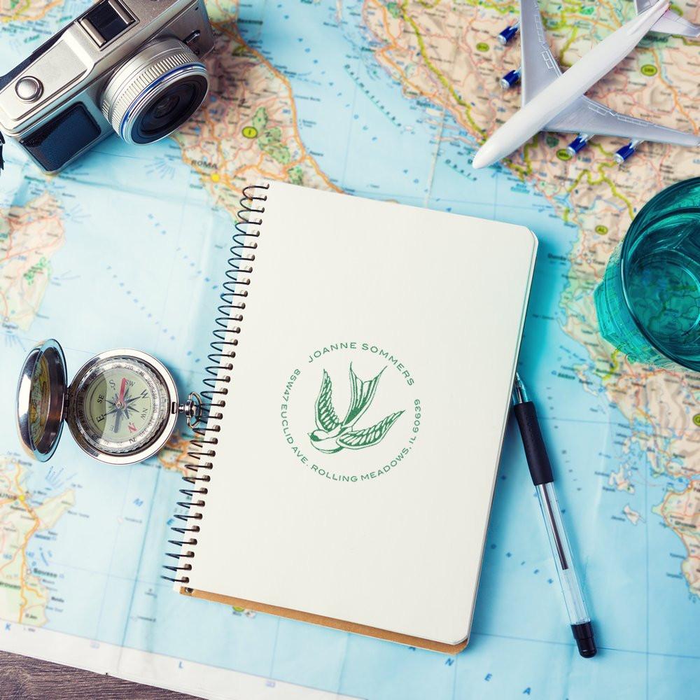 Custom return address rubber stamp on a travel journal with map, compass, camera and airplane for personalized branding.