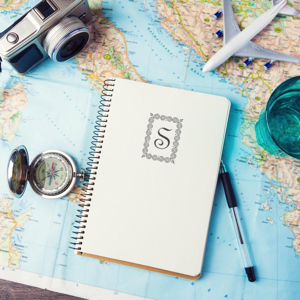 Custom rubber stamp monogram on travel journal notebook, with map, compass, camera, and airplane for travel-themed stationery.