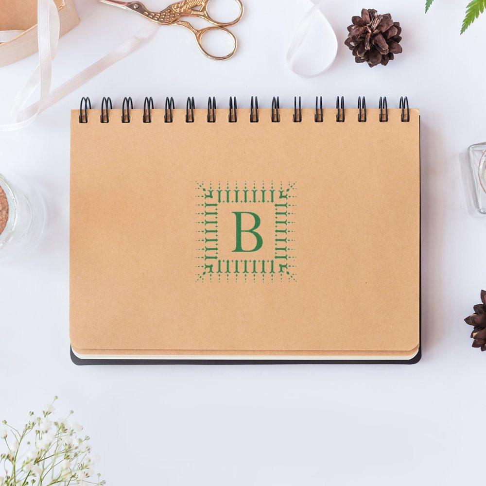 Custom monogram rubber stamp with letter B on kraft spiral notebook, ideal for personalized stationery and branding.