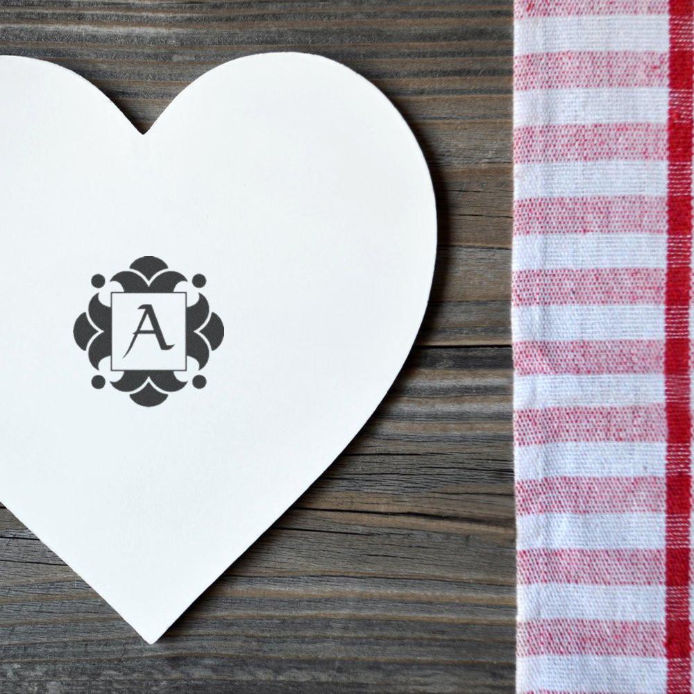 White monogram heart card with letter A on rustic wood, beside red-and-white striped fabricpersonalized wedding or party decor.