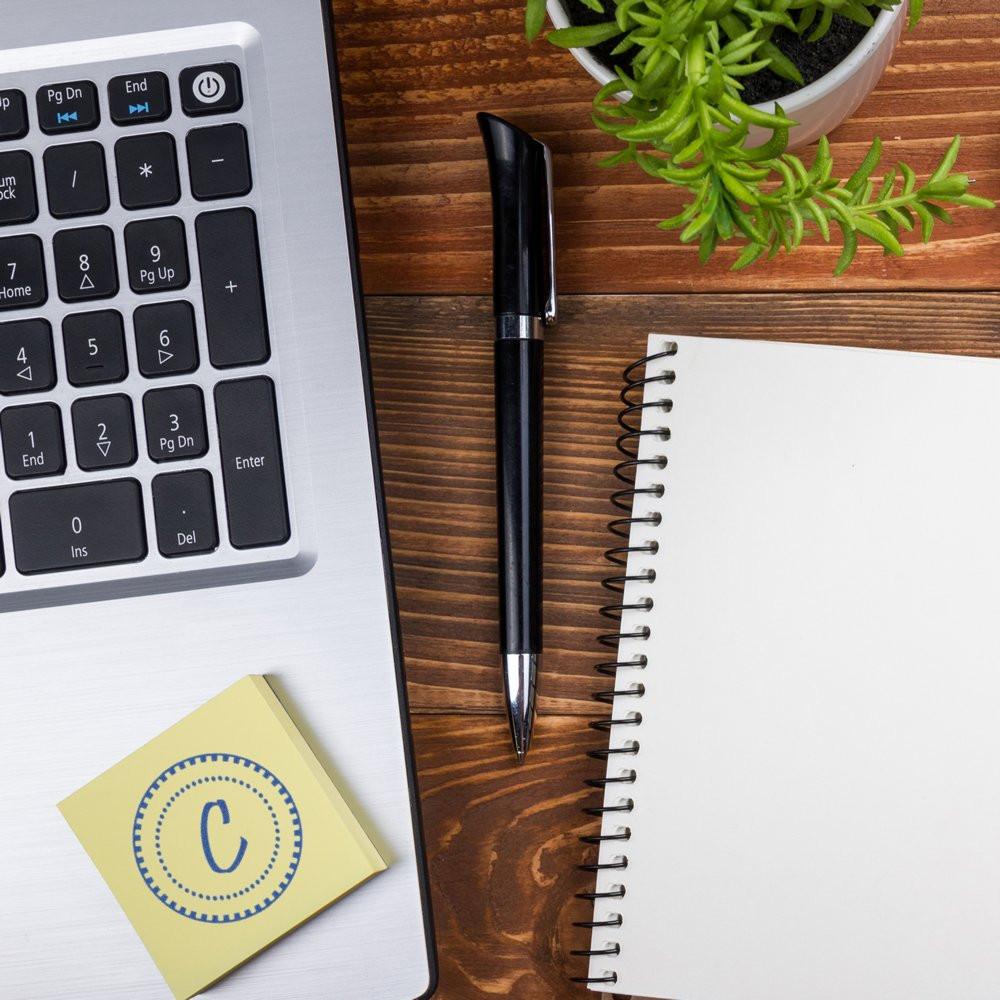 Round logo rubber stamp imprint on sticky note beside laptop, pen and notebook in a modern office workspace.
