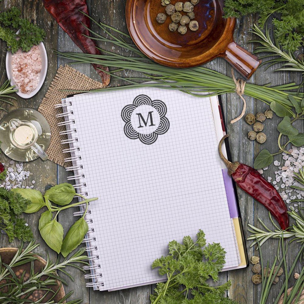 Custom floral monogram rubber stamp impression with letter M on a notebook, styled for stationery and packaging branding.