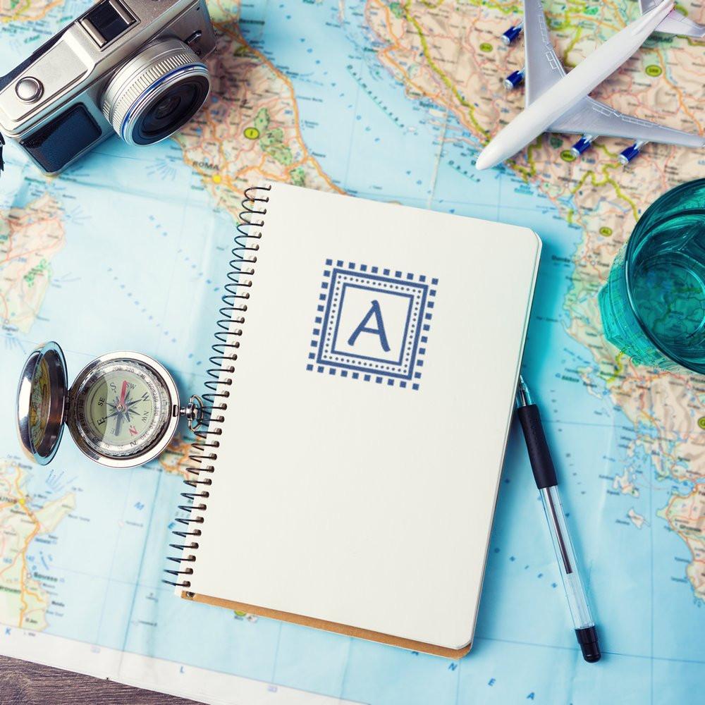 Custom travel rubber stamp design on a notebook over a world map, with camera, compass, airplane and pen for vacation planning.