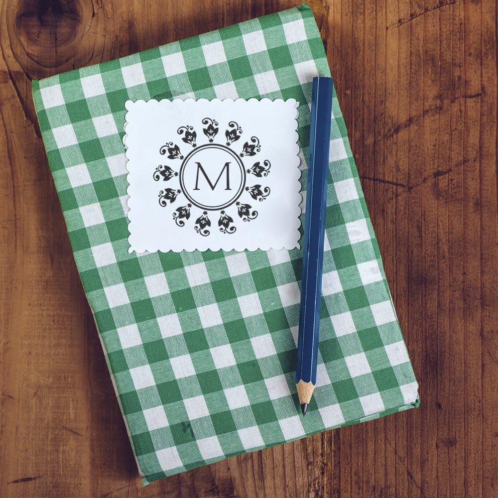 Custom monogram rubber stamp with decorative letter M on scalloped card, styled on a green gingham notebook with pencil.