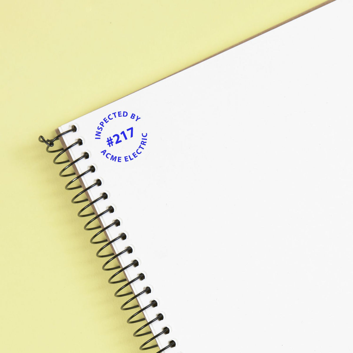 Custom blue self-inking stamp reading INSPECTED BY #217 ACME ELECTRIC on a white spiral notebook, set against a pale yellow background.