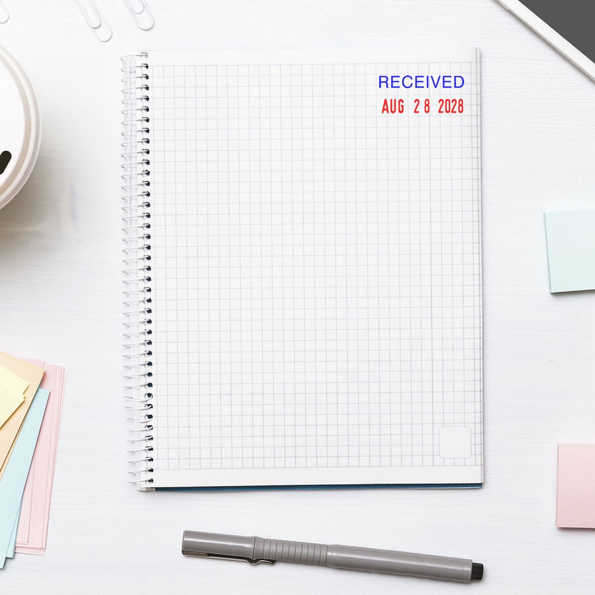 Top view of spiral grid notebook with Received Aug 28 2028 stamp, pen, and colorful sticky notes on a clean white desk.