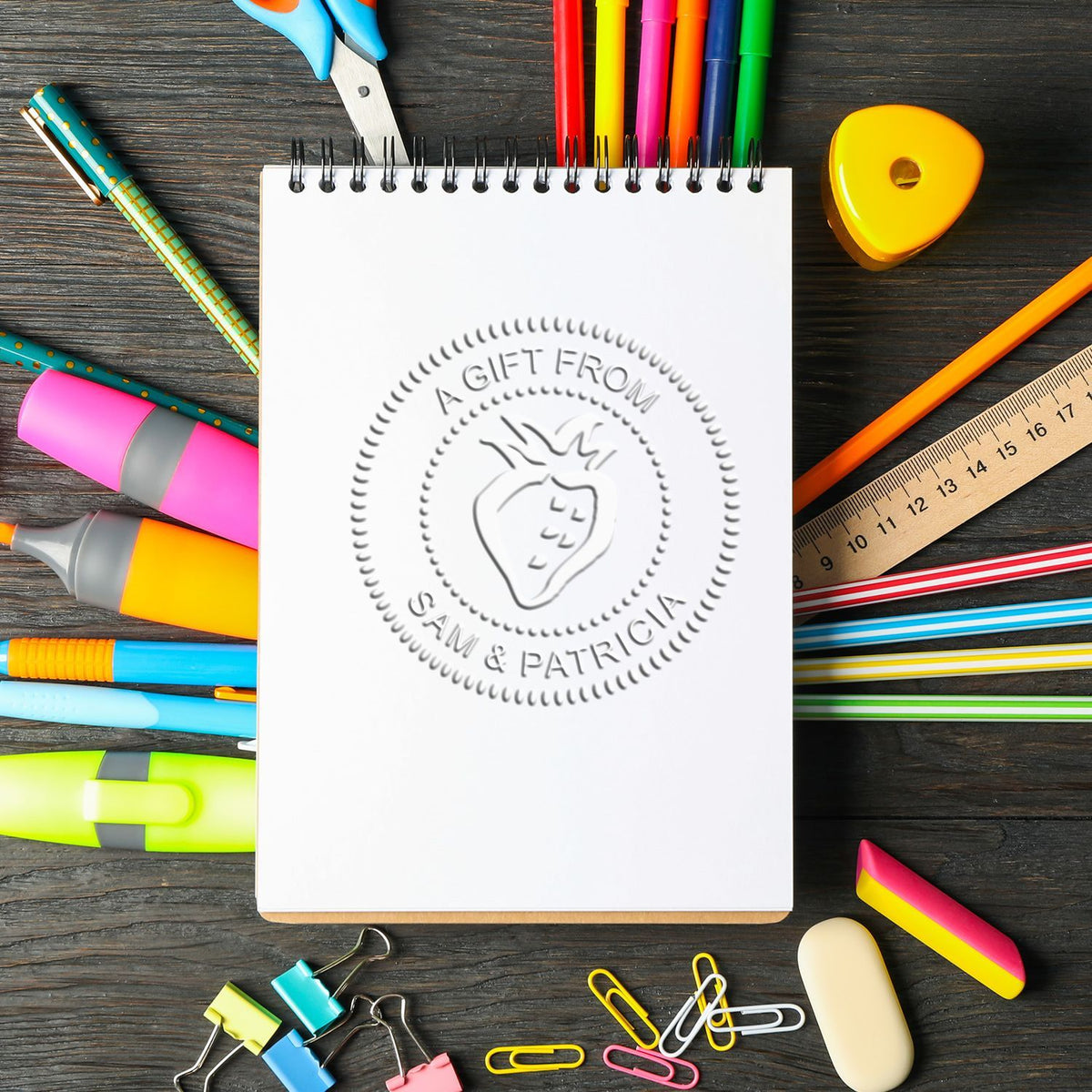 Blank spiral notebook surrounded by colorful school stationery and office supplies on a wooden desk, with a strawberry gift logo template in the center.