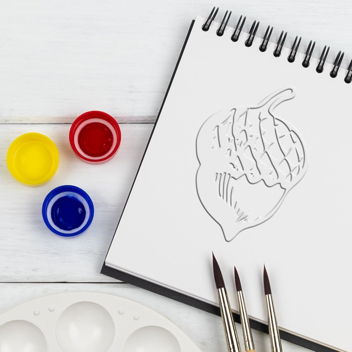 Watercolor painting supplies with sketchbook, paint pots, brushes, and a fruit outline drawing on a white wooden table.