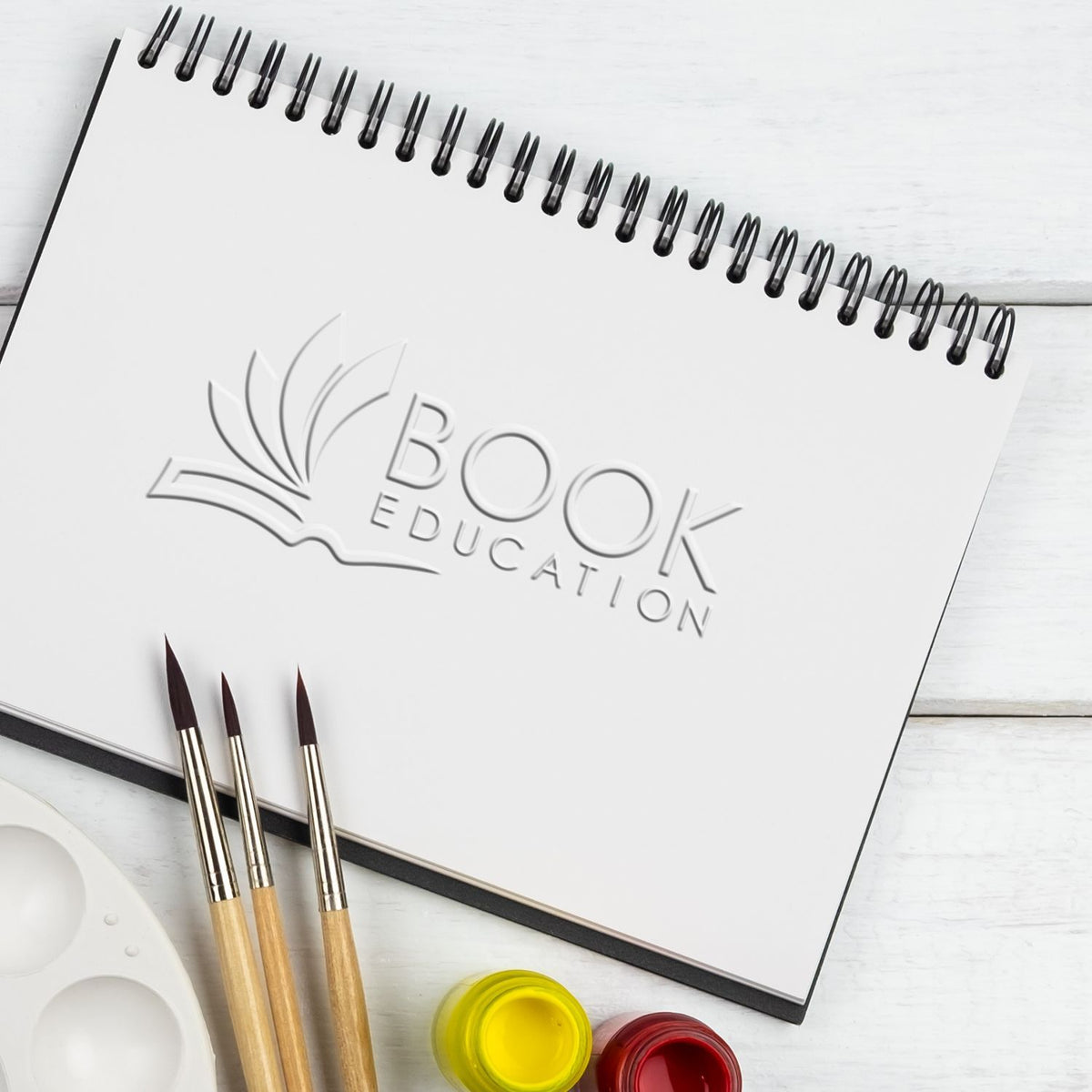 Blank spiral sketchbook with book education logo, paint brushes, palette, and paint jars on white wooden background.