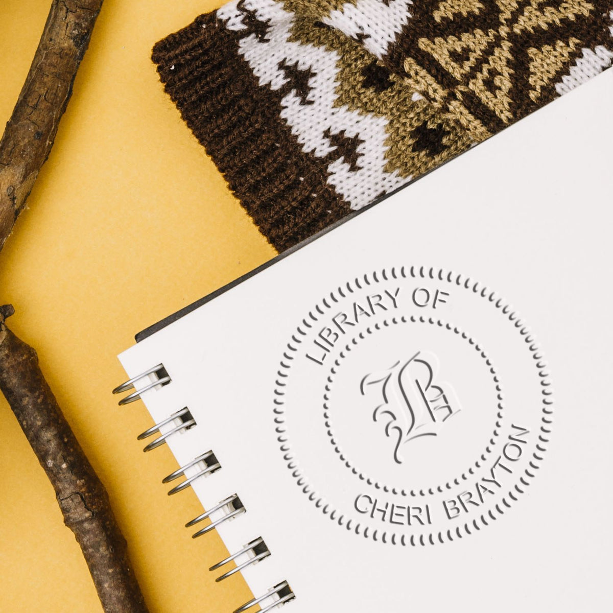 Close-up of a branded notebook with the Library of Cheri Braxton logo, knit fabric, and rustic sticks on a yellow background.