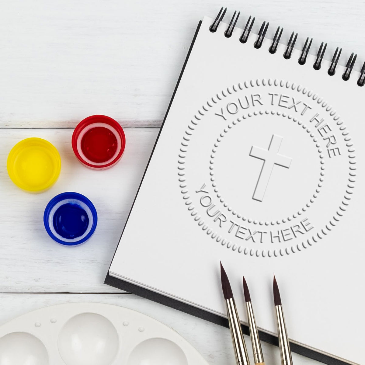 Blank spiral sketchbook mockup with embossed cross and your text here placeholder, paint brushes, color paints, and palette on a white wooden background.