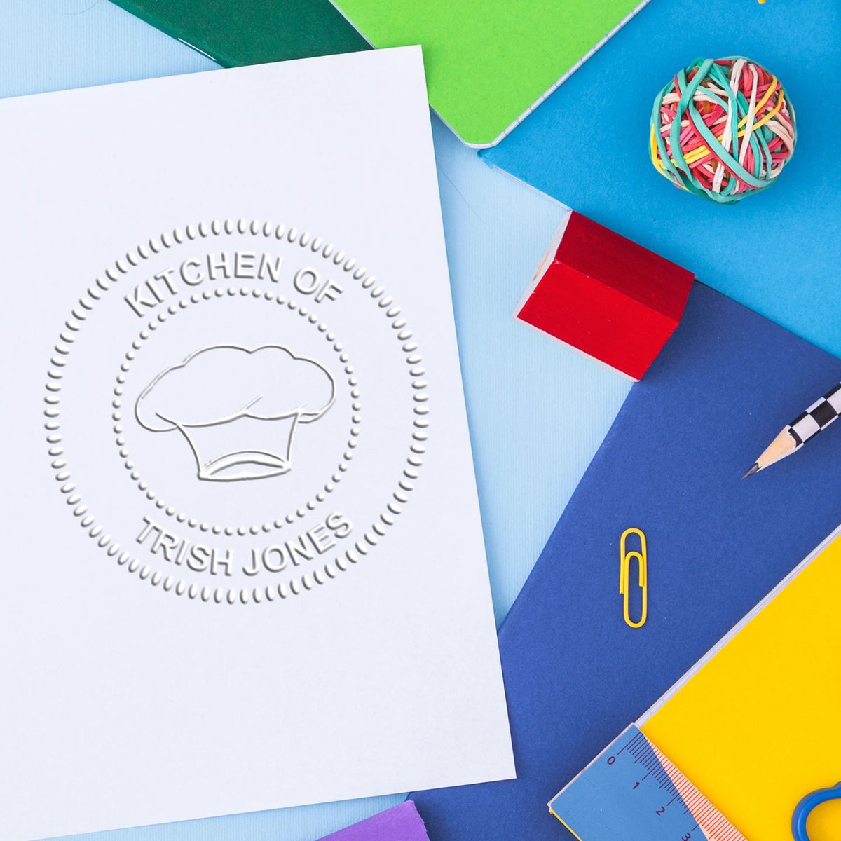 Colorful stationery flat lay with Kitchen of Trish Jones notebook, pencil, paper clips, and office supplies on a blue background