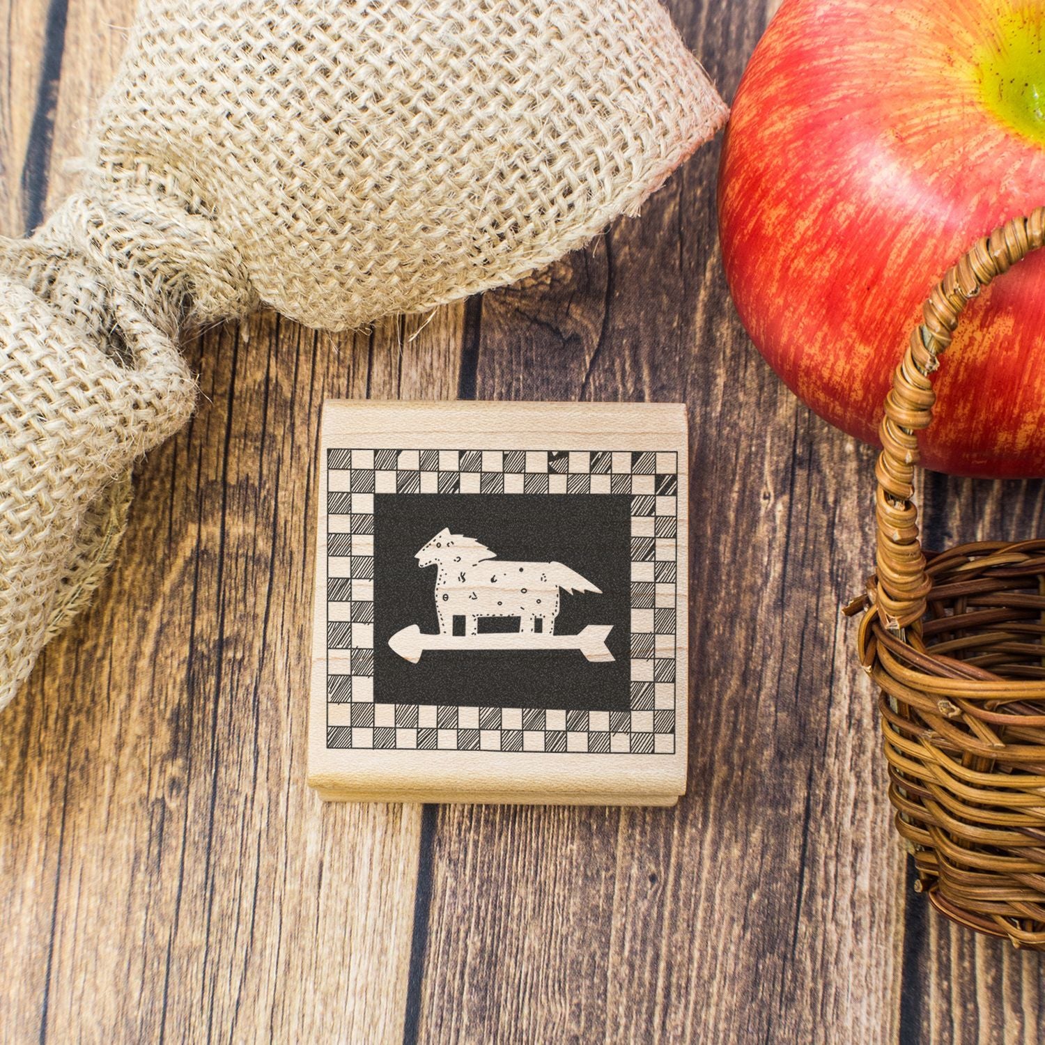 Rustic horse stamp beside apple, burlap, and wicker basket on a wooden table, autumn farmhouse decor.